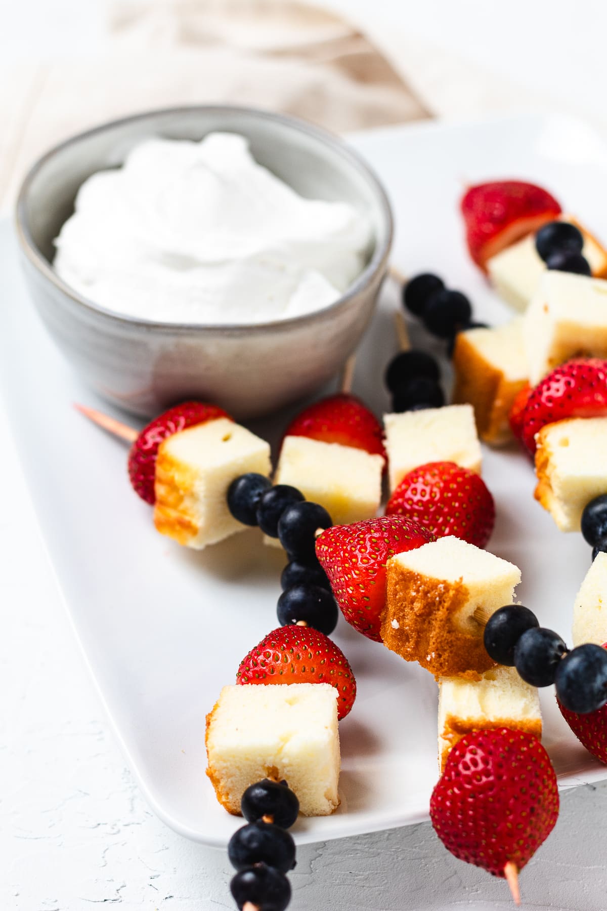 Patriotic, red, white, and blue pound cake skewers on a white platter with a bowl of whipped cream.