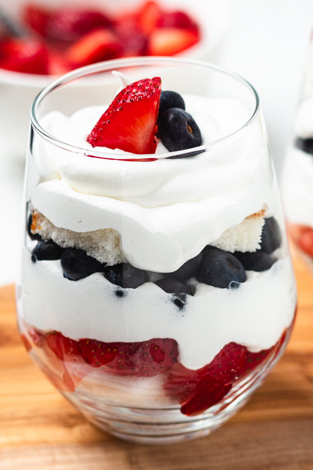 Layered strawberry, blueberry, cake, and whipped cream trifles in glass cups on a wooden board.