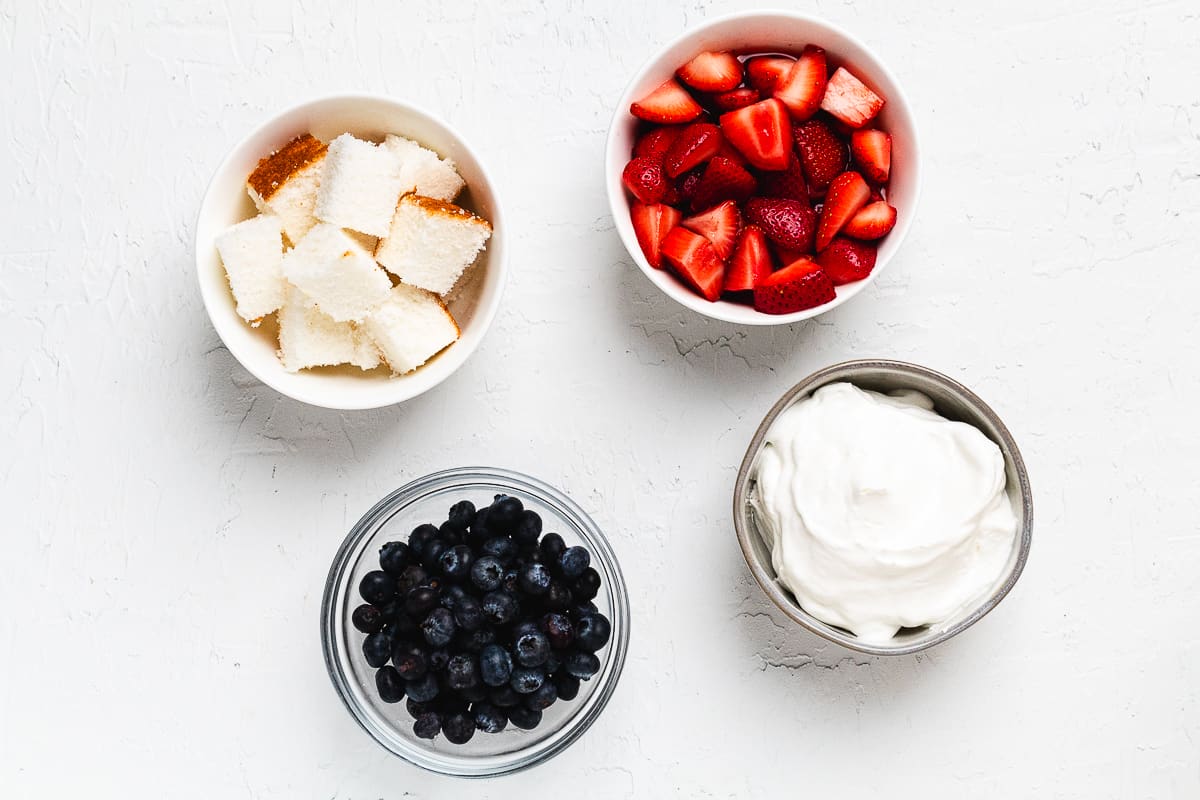 Bowls of cubed angel food cake, sliced strawberries, blueberries, and whipped cream.