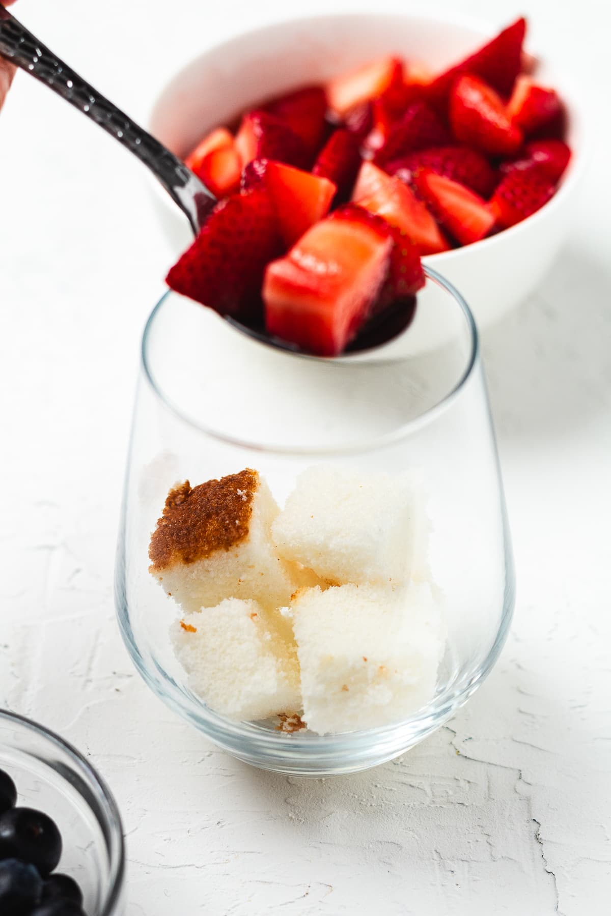 Hand spooning strawberries into a glass filled with cubed angel food cake.