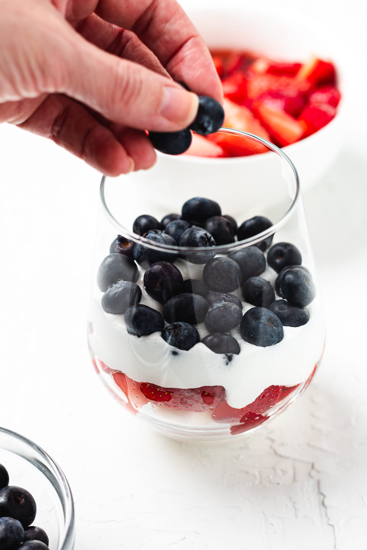 Hand adding blueberries to a glass full of cubed angel food cake and whipped cream.