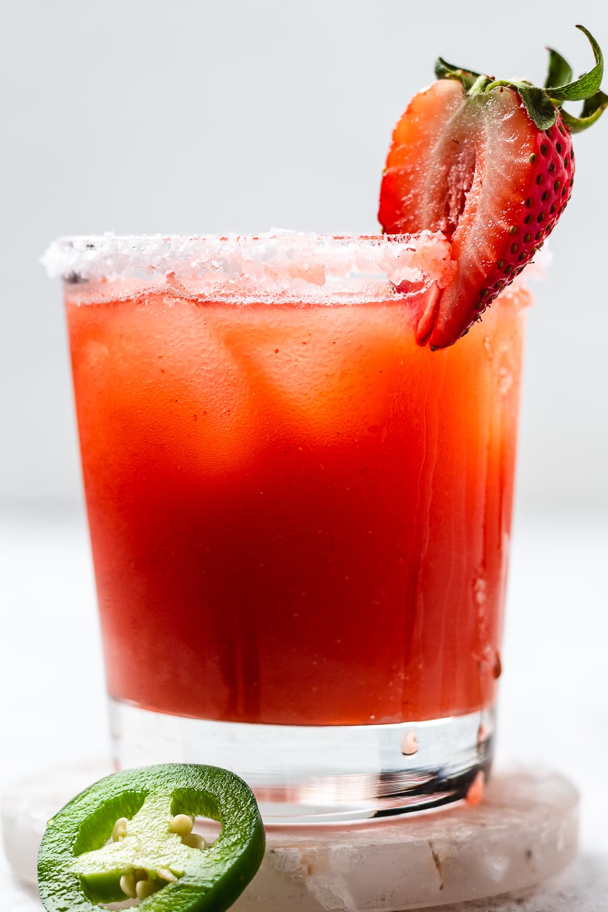 Glass of red orange drink with ice and a salted rim topped with a strawberry and a jalapeรฑo slice at the base.