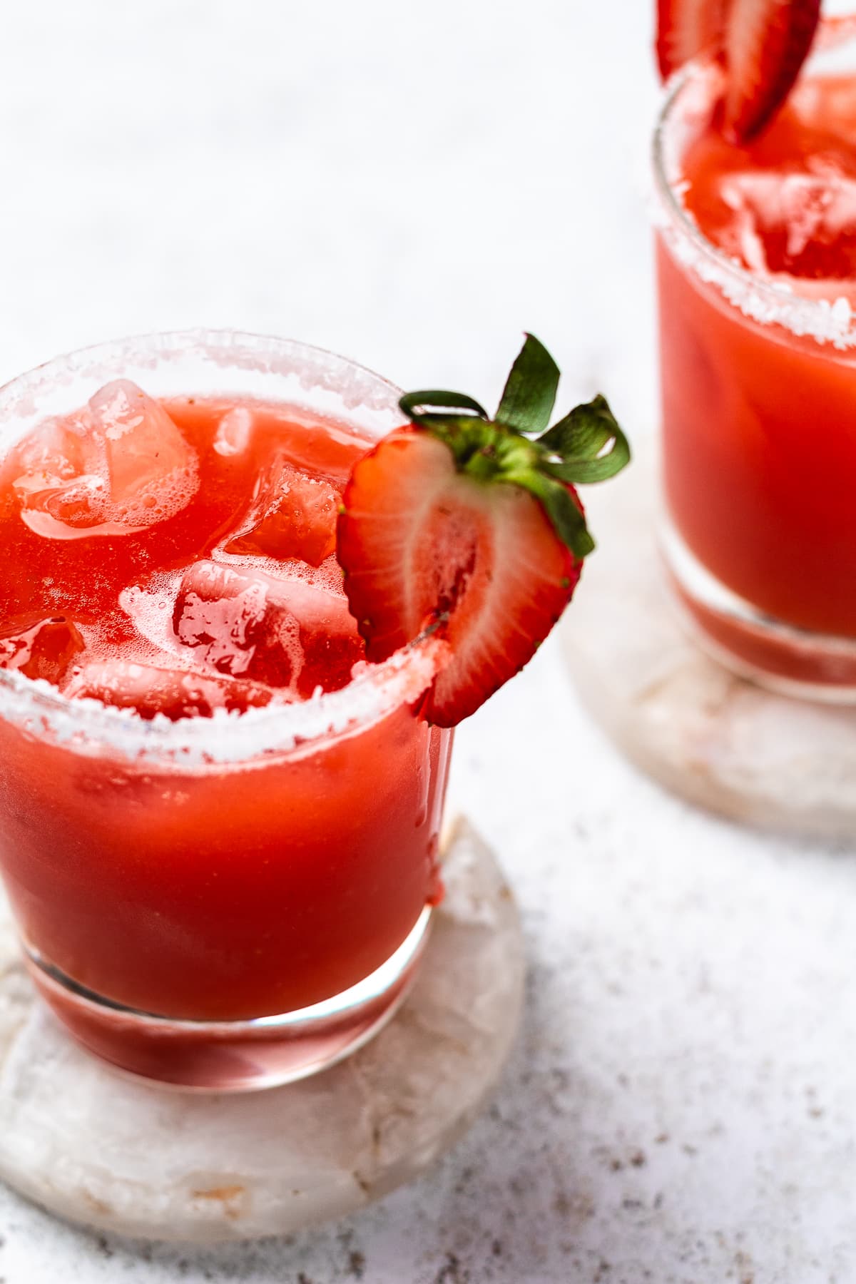 Two glasses of red drink with ice and strawberry slices on salted rims on stone coasters.