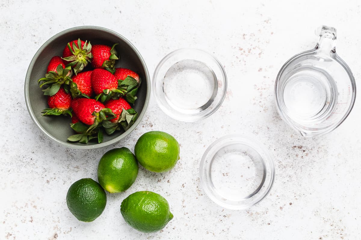 Bowl of strawberries with limes and three clear containers of water arranged on a light surface.