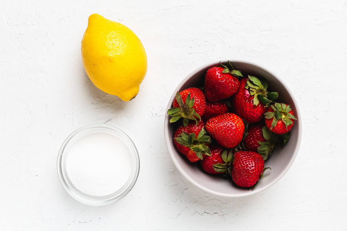 Ingredients for strawberry topping including whole strawberries, lemon, and sugar.