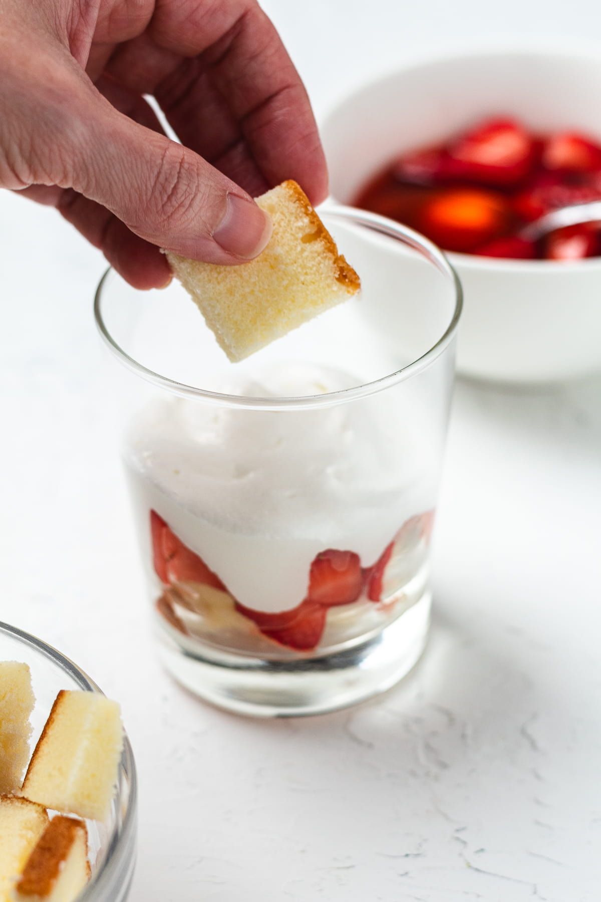Placing cake cubes into a glass over whipped cream to assemble layered strawberry trifle cups.