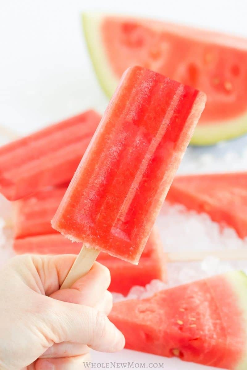 Hand holding watermelon popsicle with fresh watermelon slices in background.