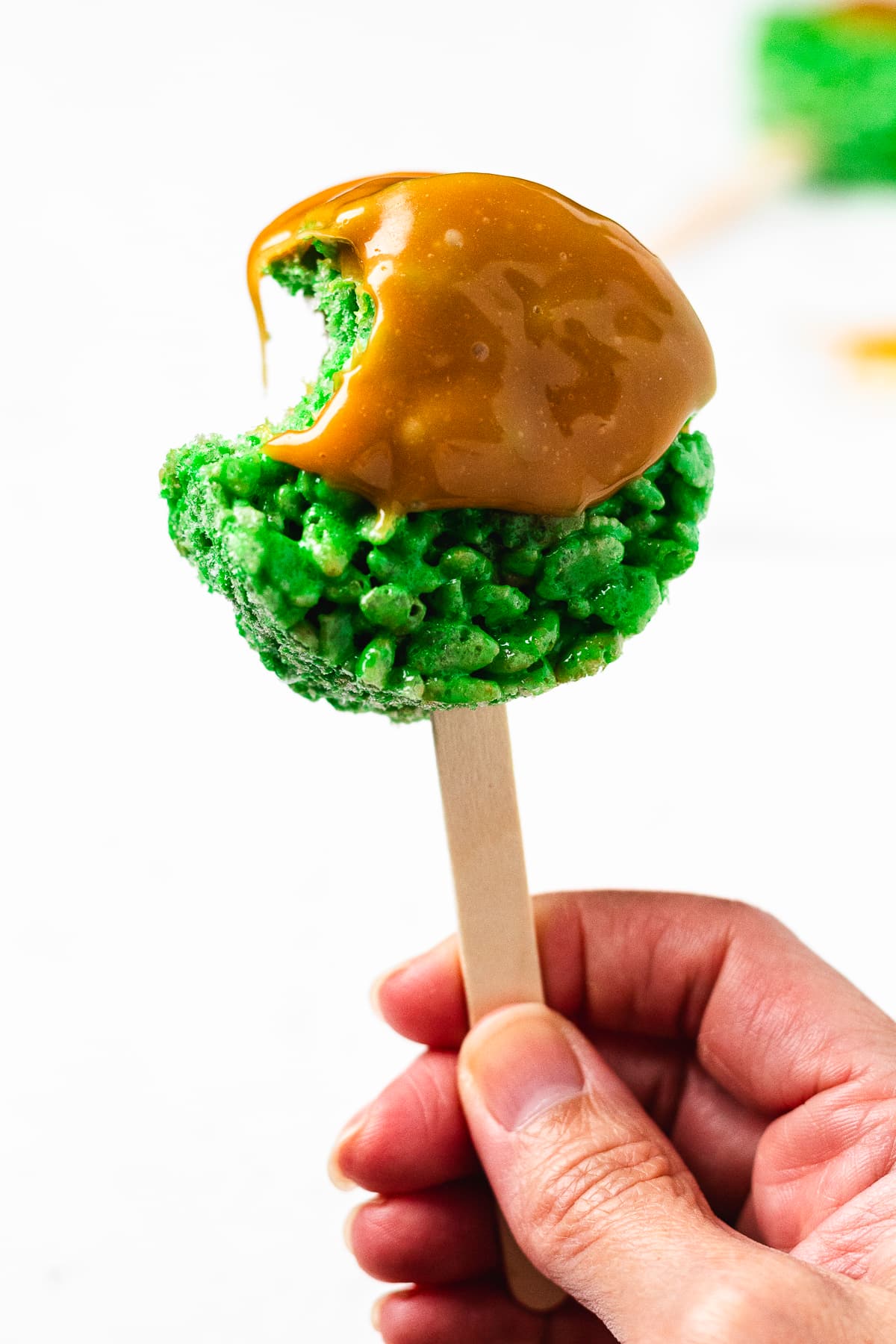 Bitten caramel apple Rice Krispie treat pop held by the stick.