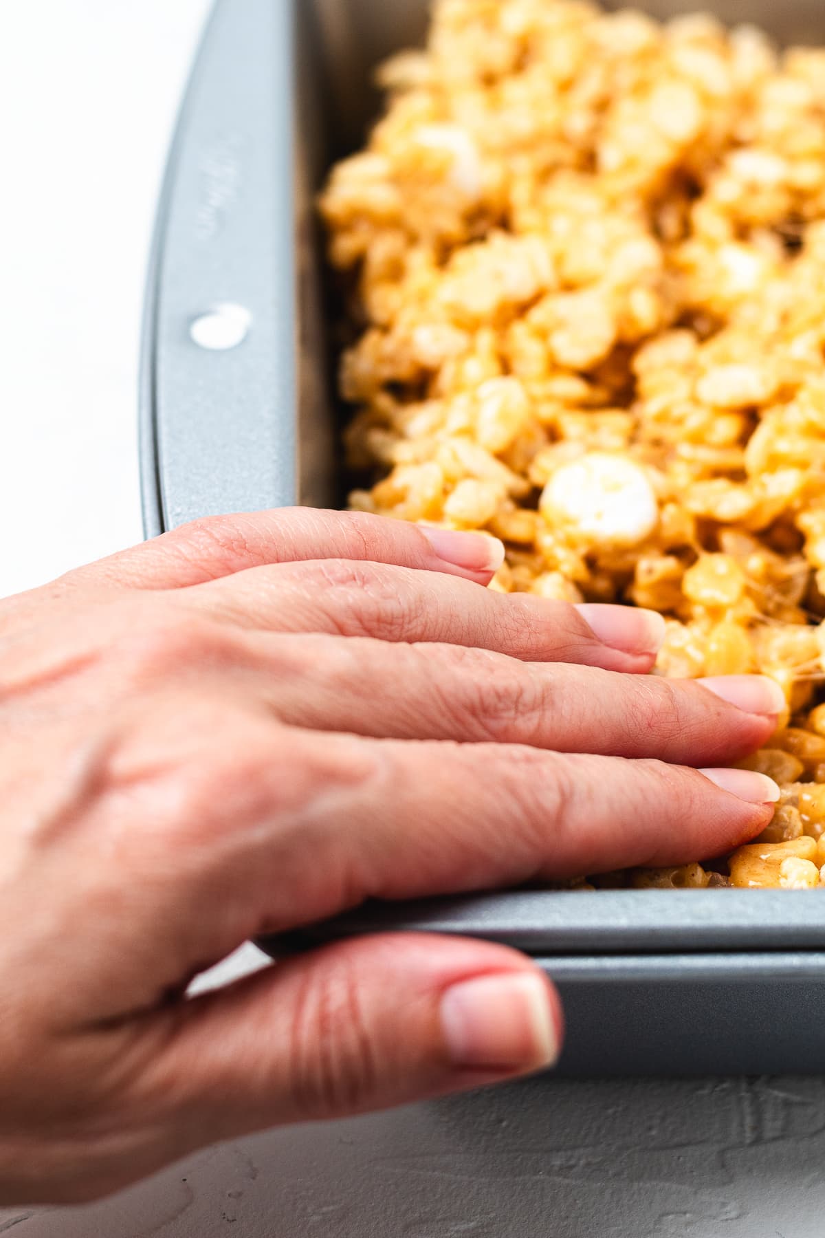 Hand gently pressing peanut butter Rice Krispie treat mixture into a metal baking pan.