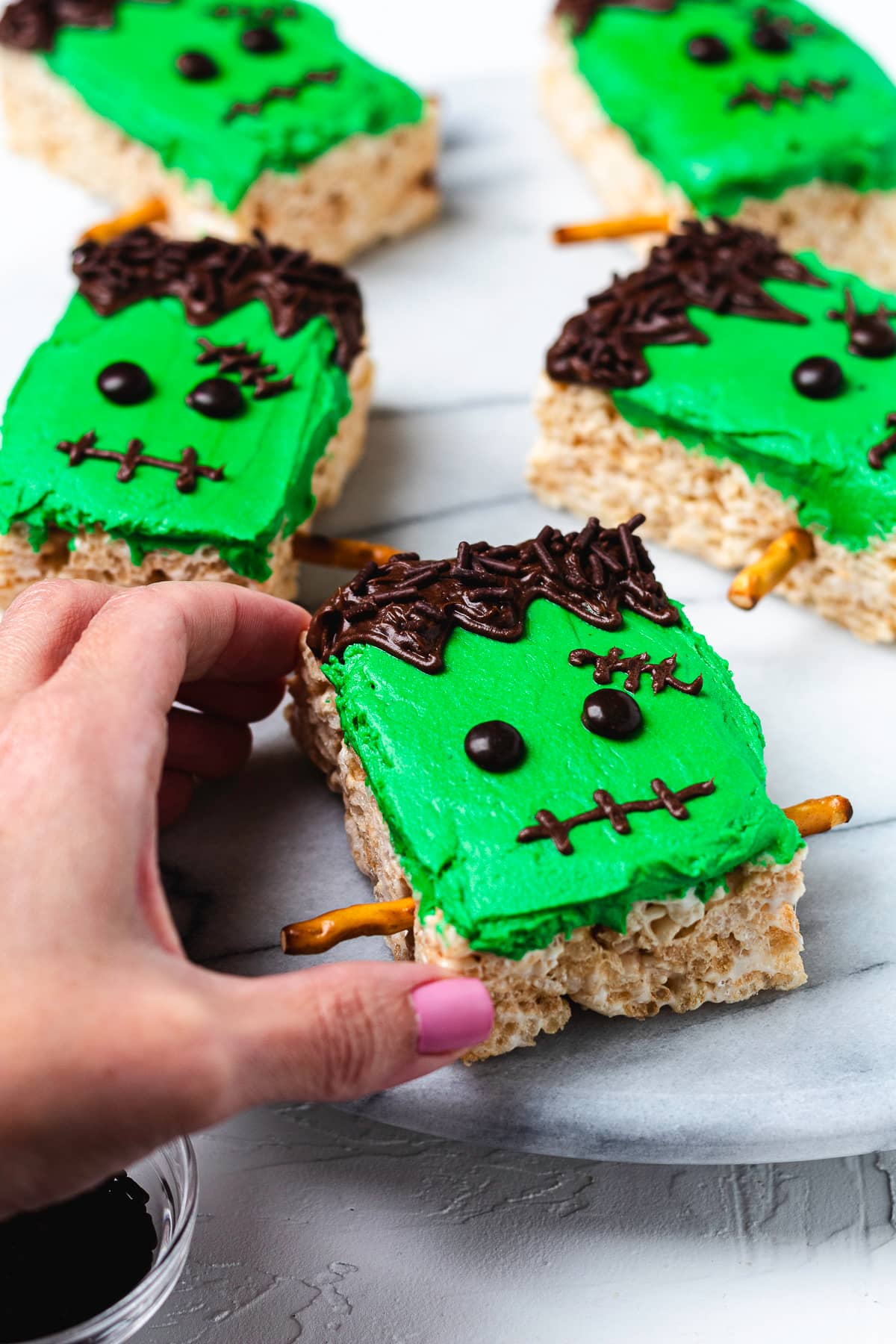 Hand holding decorated Frankenstein Rice Krispie treat with green frosting and pretzel bolts.