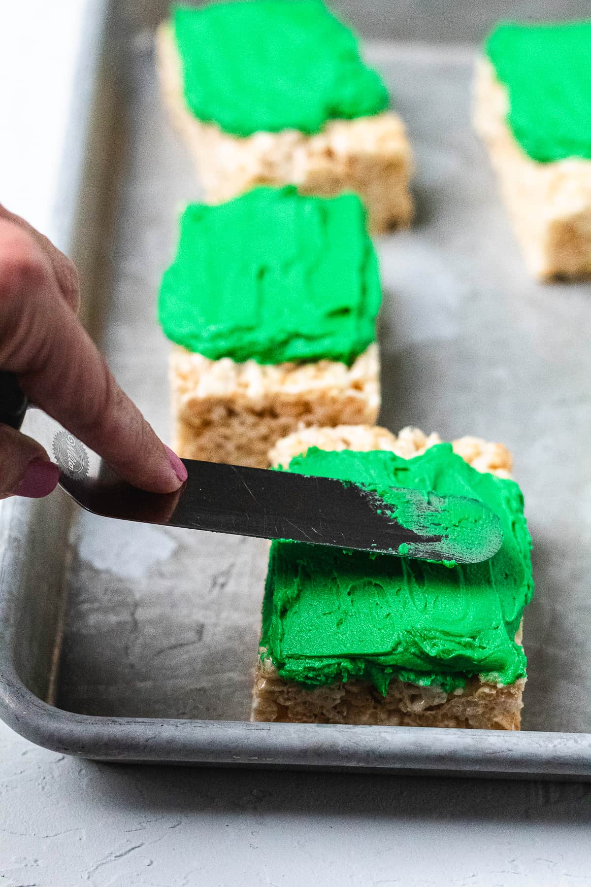 Spreading green frosting evenly over Rice Krispie treat with an offset spatula.