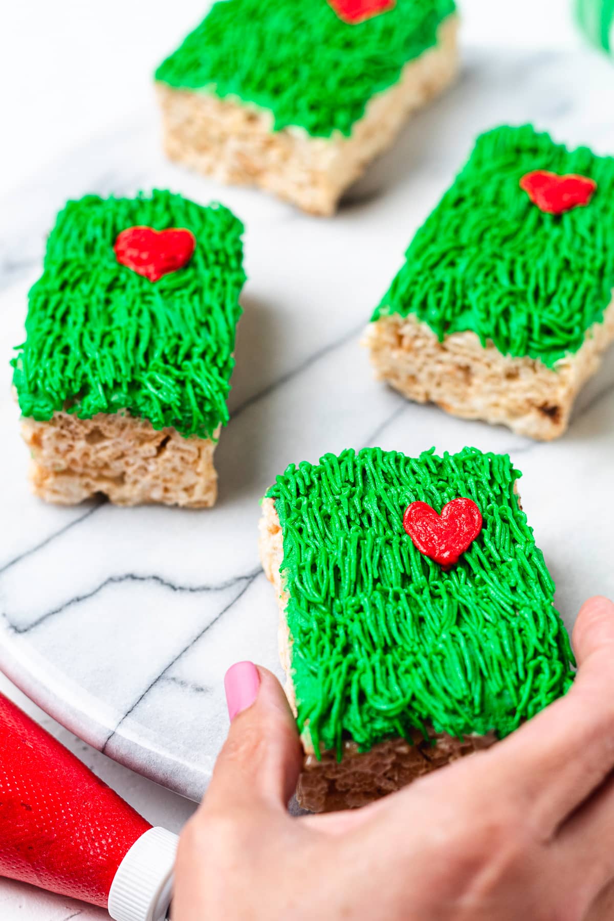 Hand holding a Grinch Rice Krispie treat with green frosting and a red heart.