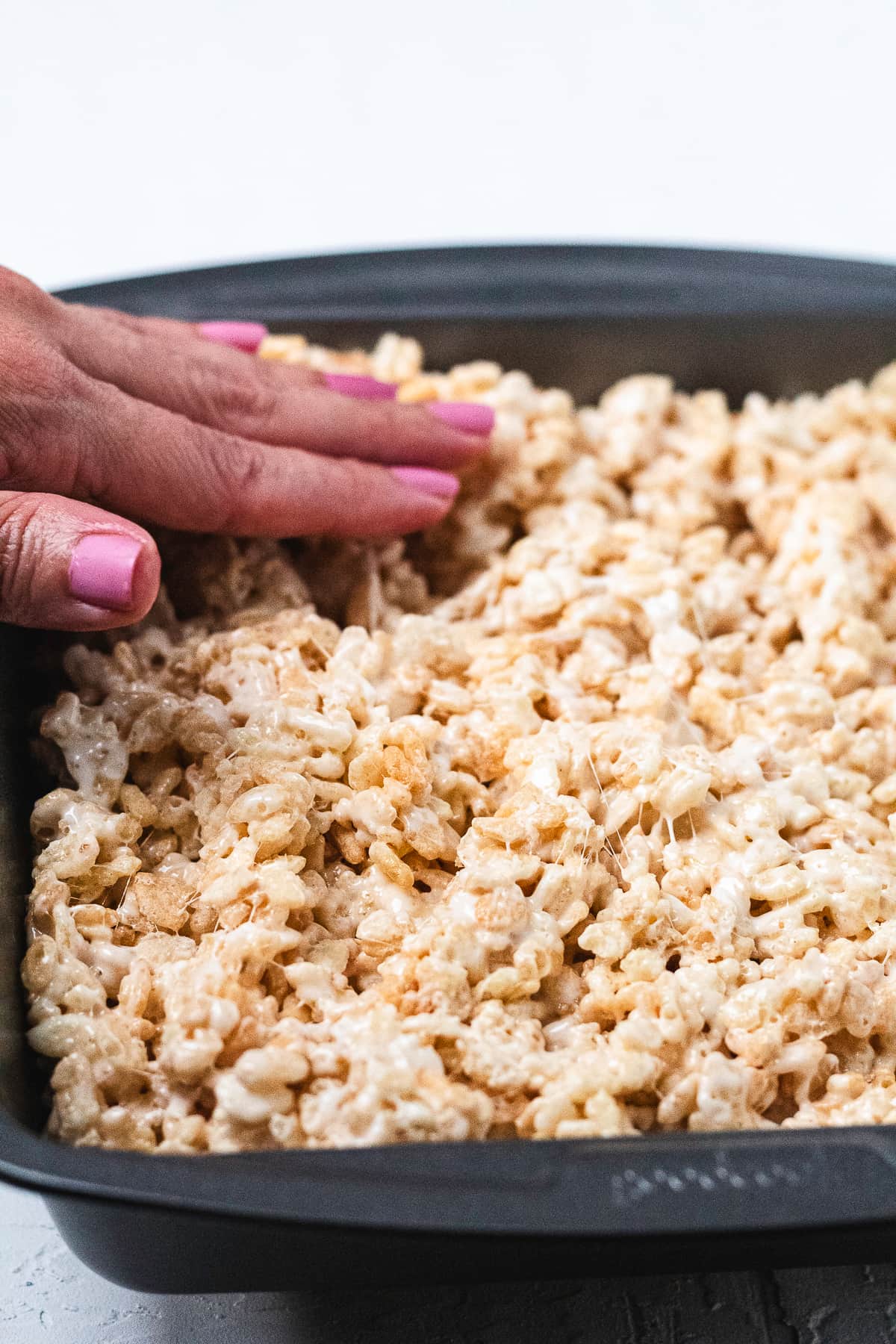Hand patting down Rice Krispie treats mixture in a dark gray pan.