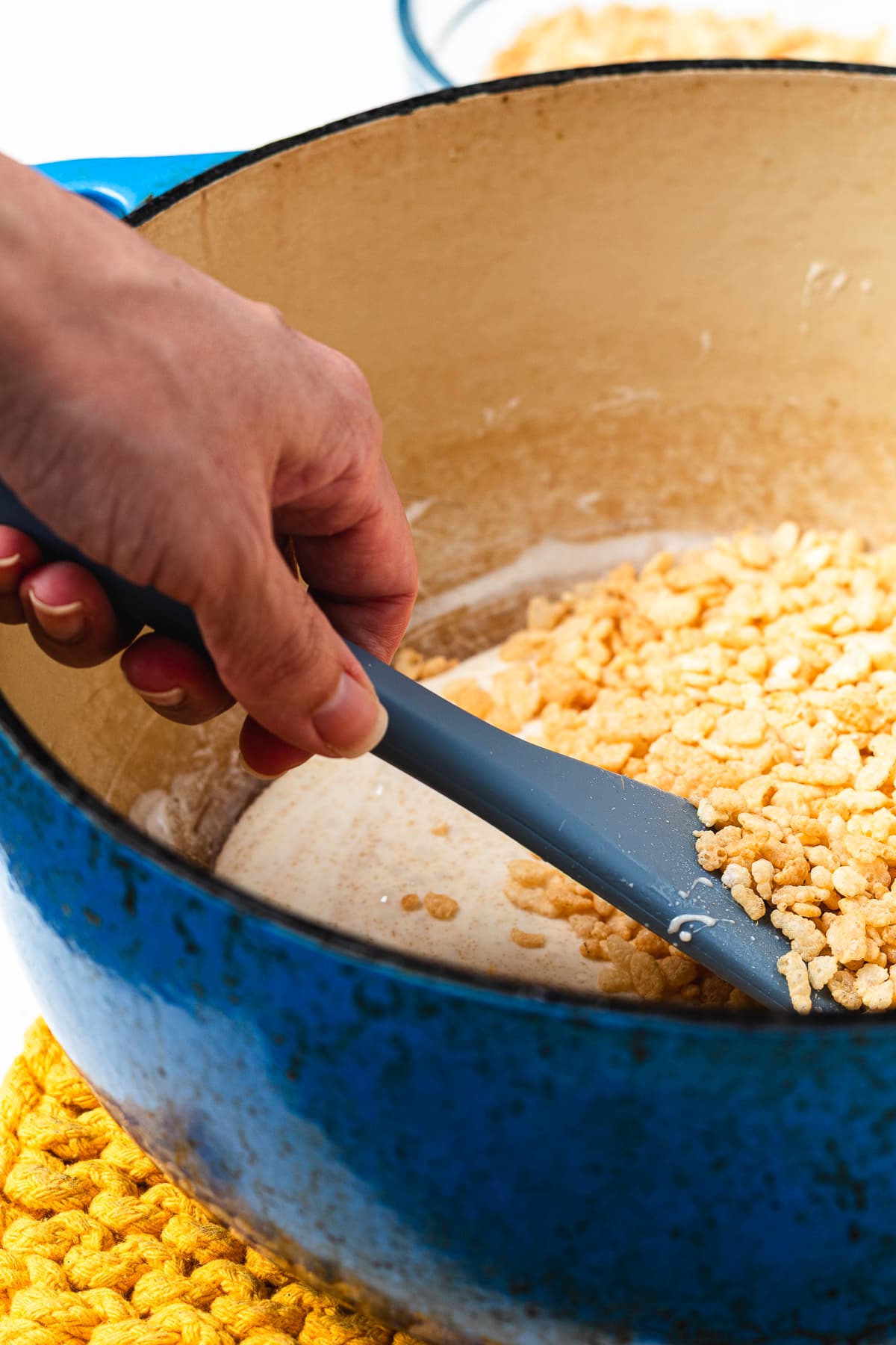 Rice Krispie cereal being stirred into the melted marshmallow mixture in a blue Dutch oven.
