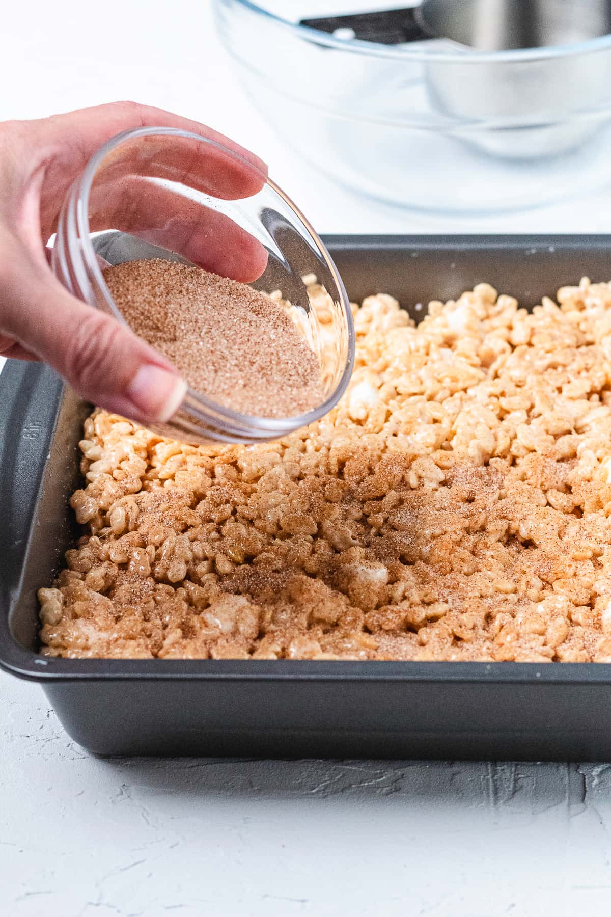 Cinnamon sugar being sprinkled over Snickerdoodle Rice Krispie Treats in a baking pan.