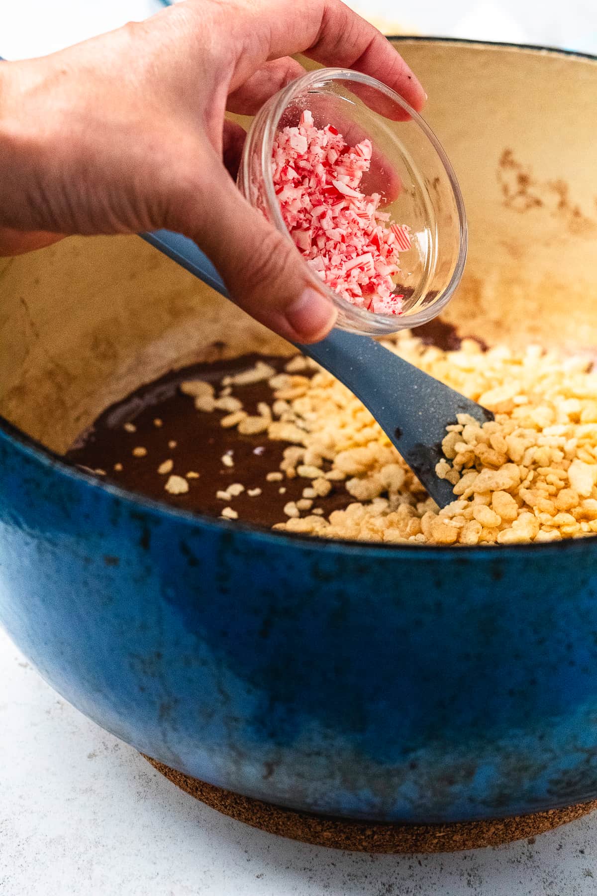 A hand holds crushed candy canes over a pot of chocolate Rice Krispie treats while mixing.