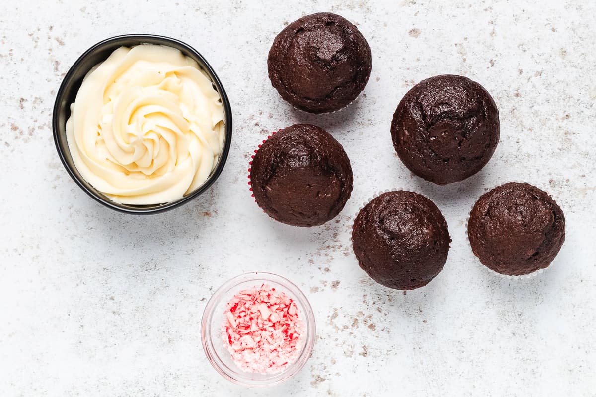Chocolate cupcakes arranged beside bowls of creamy peppermint frosting and crushed candy canes.