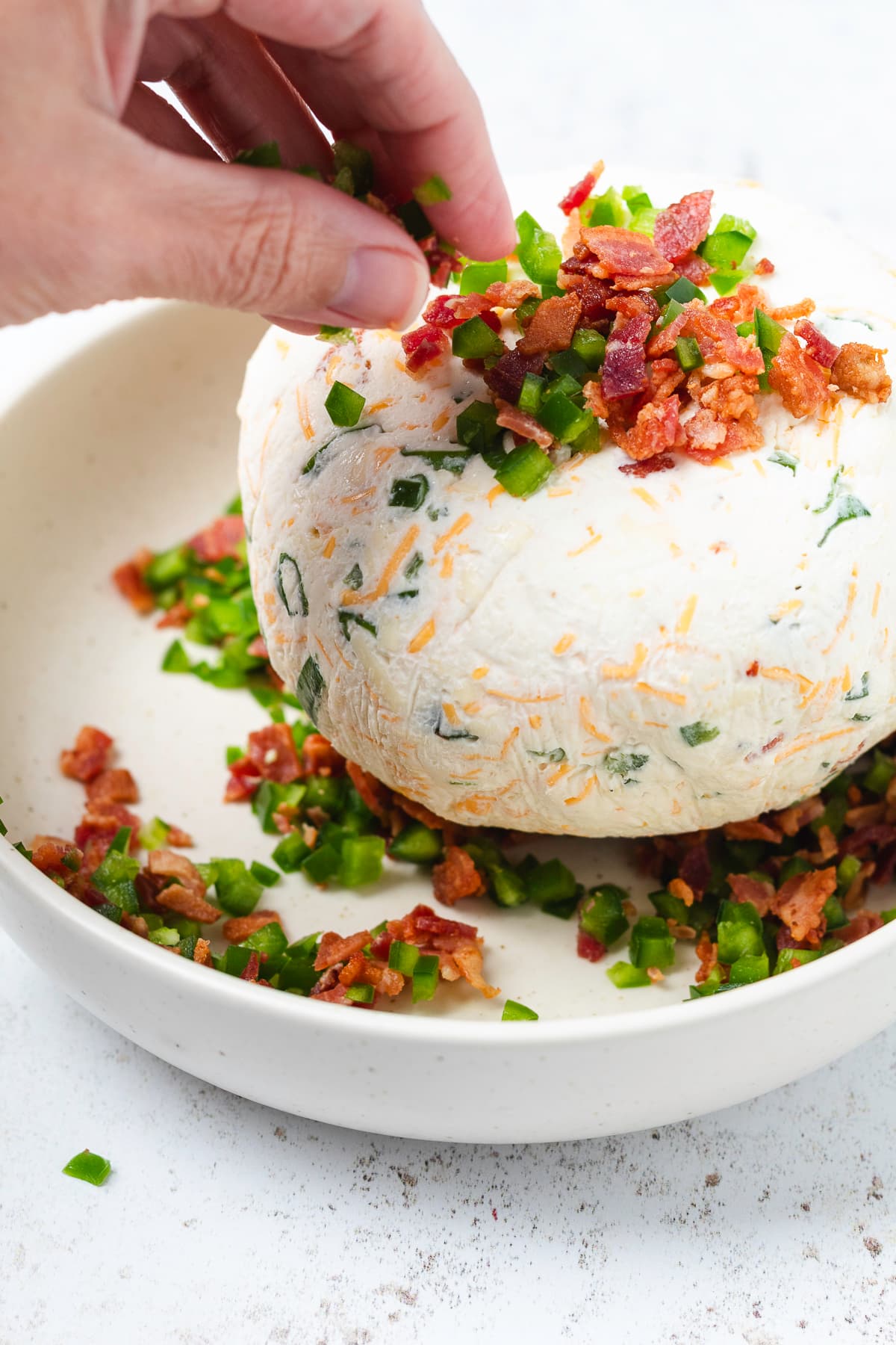 A hand adds chopped jalapeños and crispy bacon to the outside of a cream cheese ball.