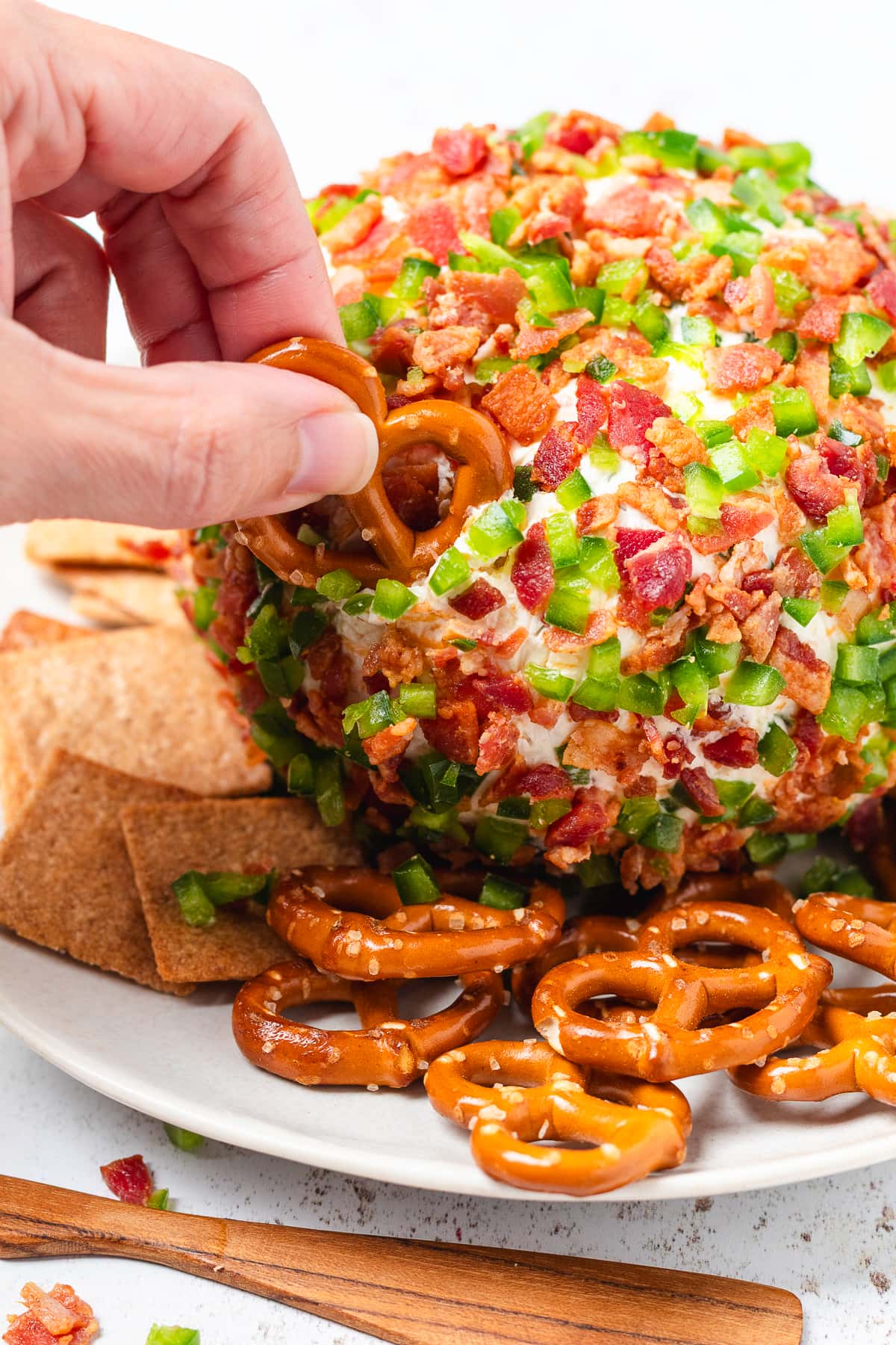 A hand dips a pretzel into a bacon jalapeño cheese ball coated with crispy bacon and diced jalapeños.