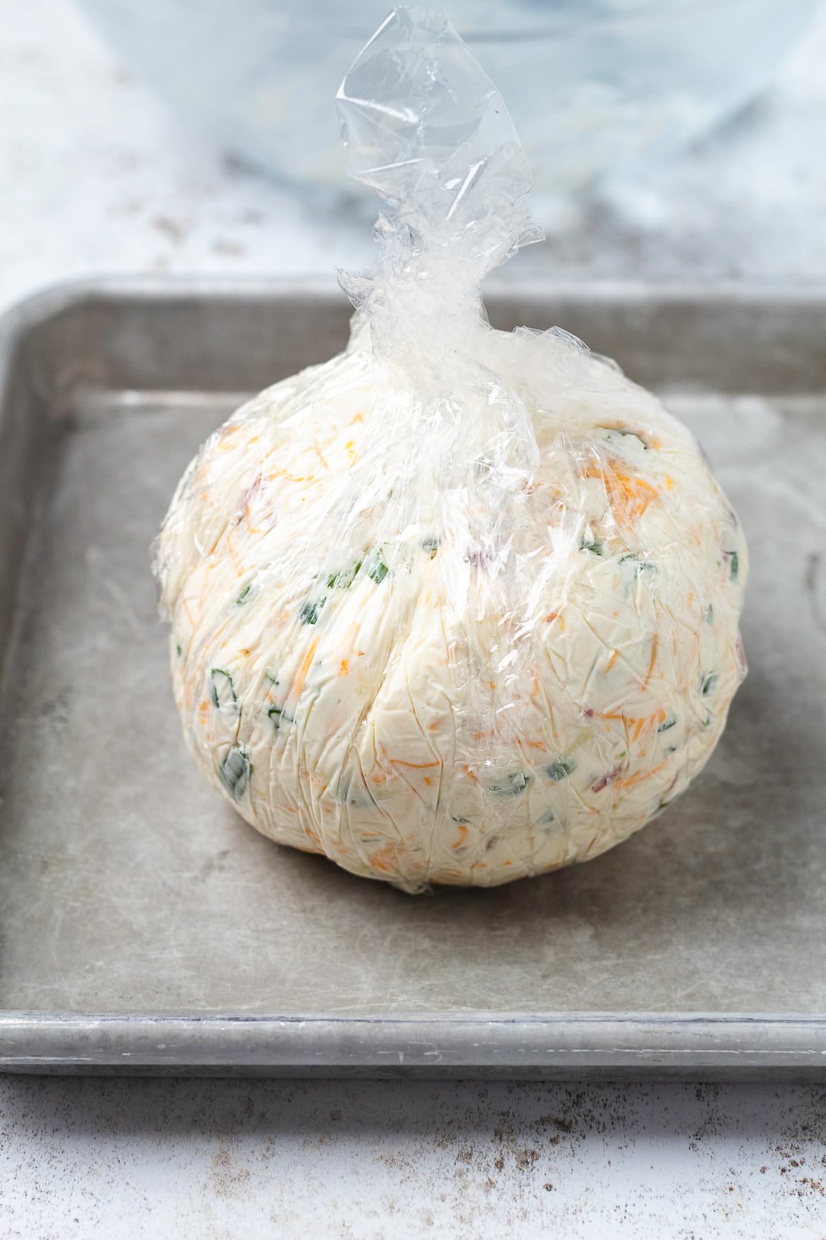 Homemade bacon jalapeño cheese ball wrapped tightly in plastic wrap on a baking sheet.