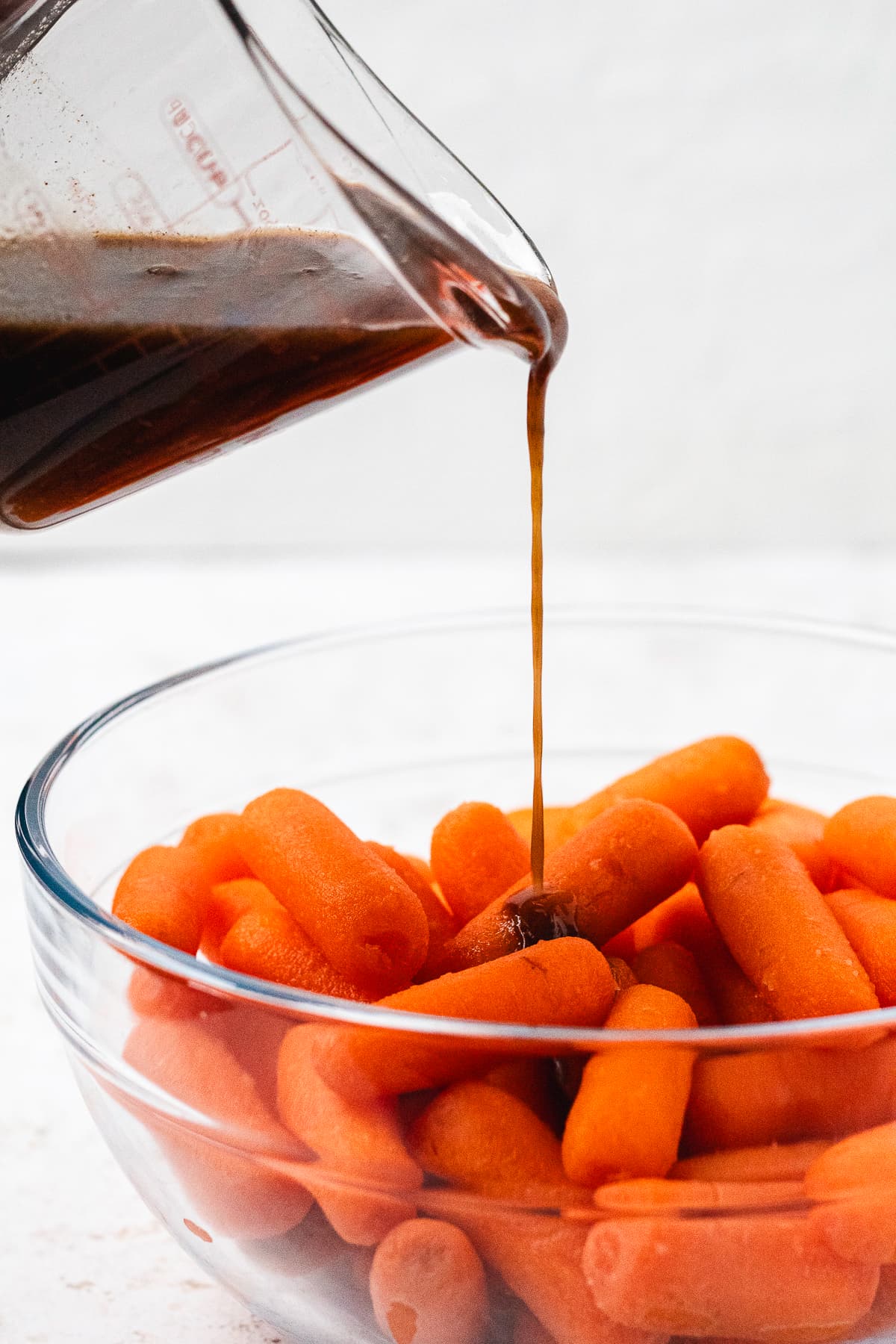 Brown sugar glaze being poured from a glass measuring cup over baby carrots in a mixing bowl.