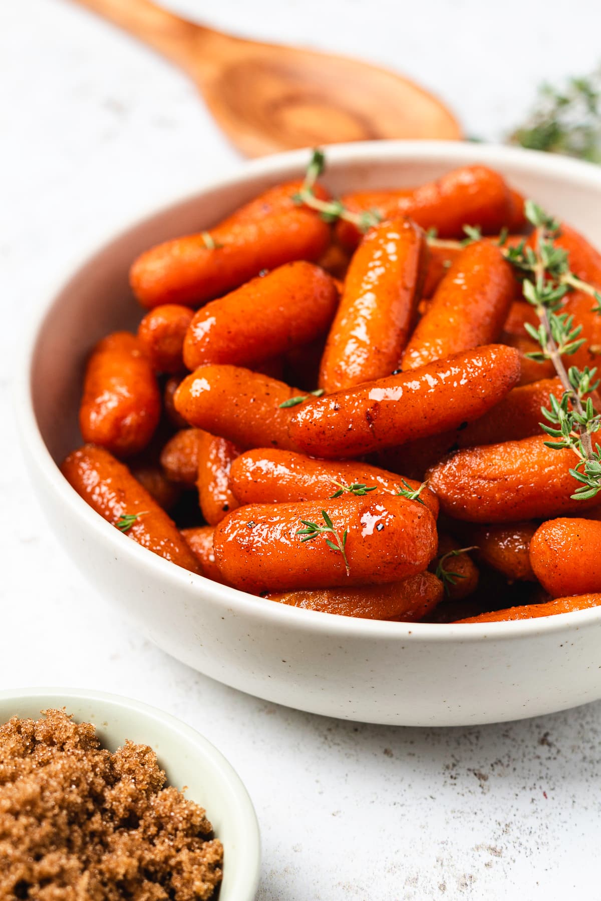 Glossy brown sugar glazed baby carrots in a white bowl, garnished with fresh thyme, with brown sugar in the foreground.
