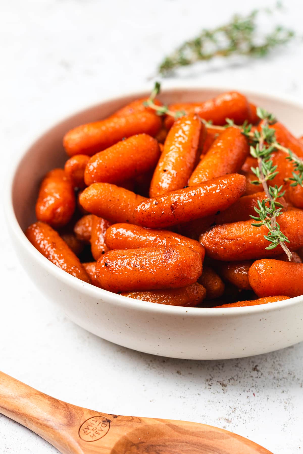 Close-up of brown sugar glazed baby carrots topped with fresh thyme in a white ceramic serving bowl.