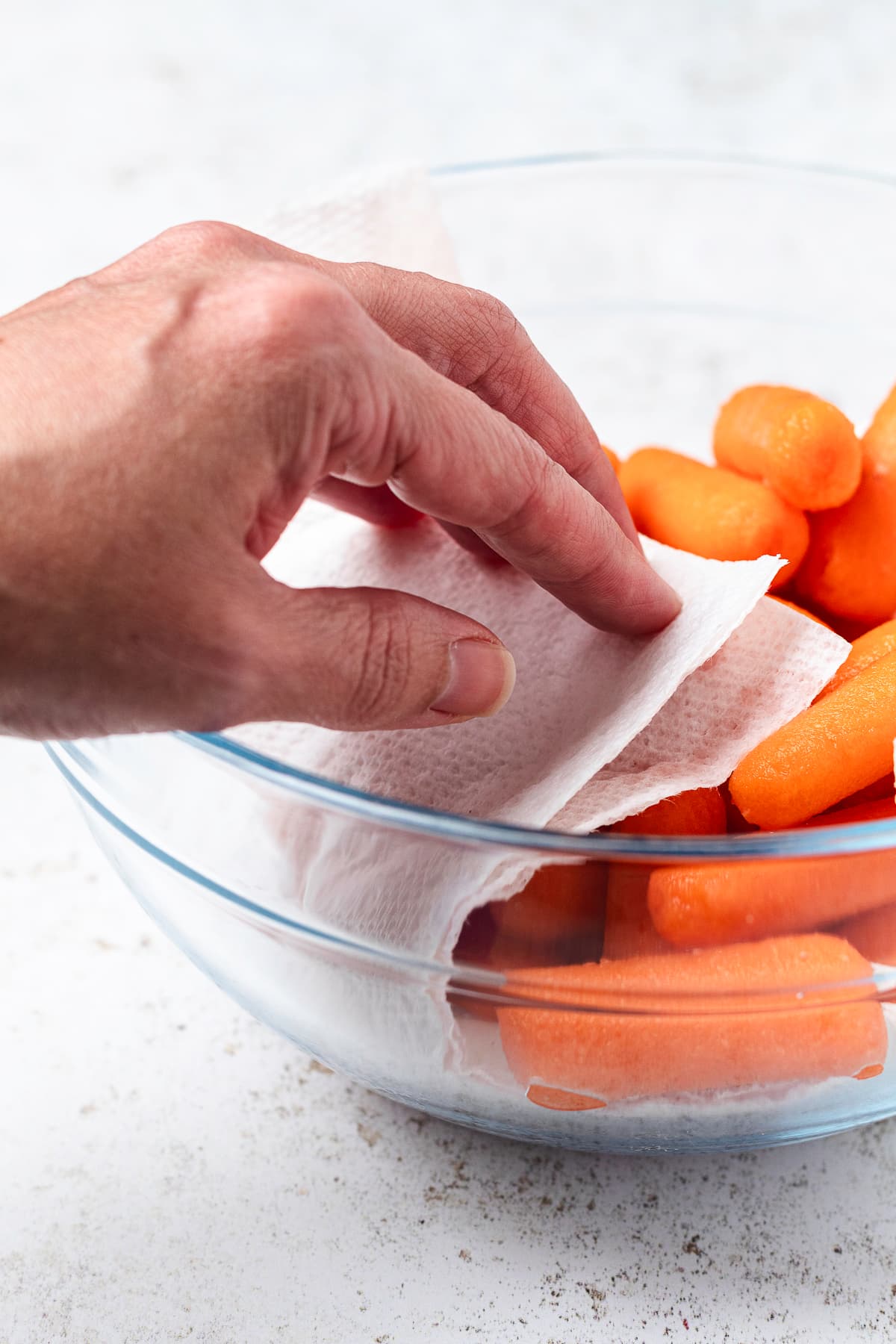 A hand patting baby carrots dry with a paper towel in a clear glass mixing bowl.