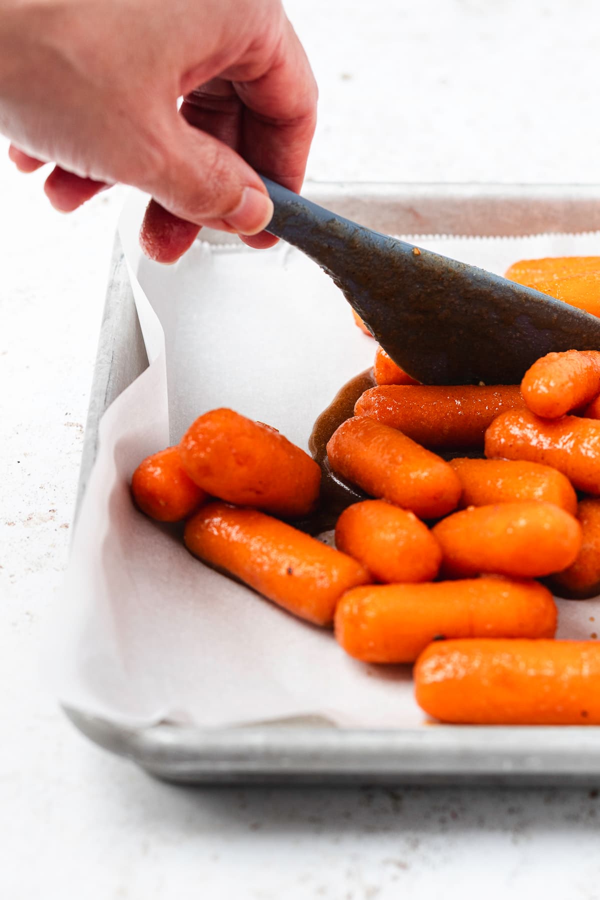 A hand using a spatula to spread glazed baby carrots onto a parchment-lined baking sheet.