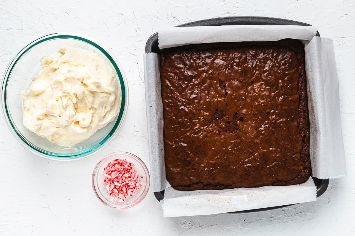 Baked chocolate brownies in a square pan beside bowls of peppermint frosting and crushed candy canes.