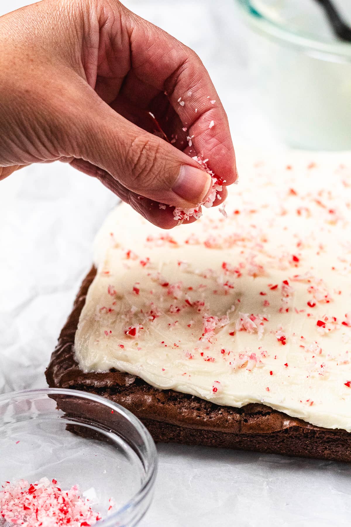 Crushed candy canes being sprinkled over creamy peppermint frosting on a pan of chocolate brownies.