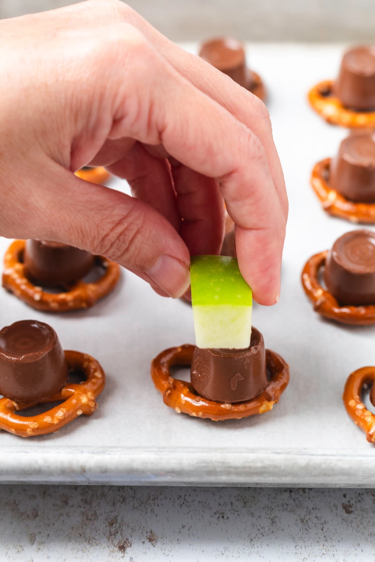 A hand presses a piece of green apple into a softened Rolo candy on a mini pretzel.