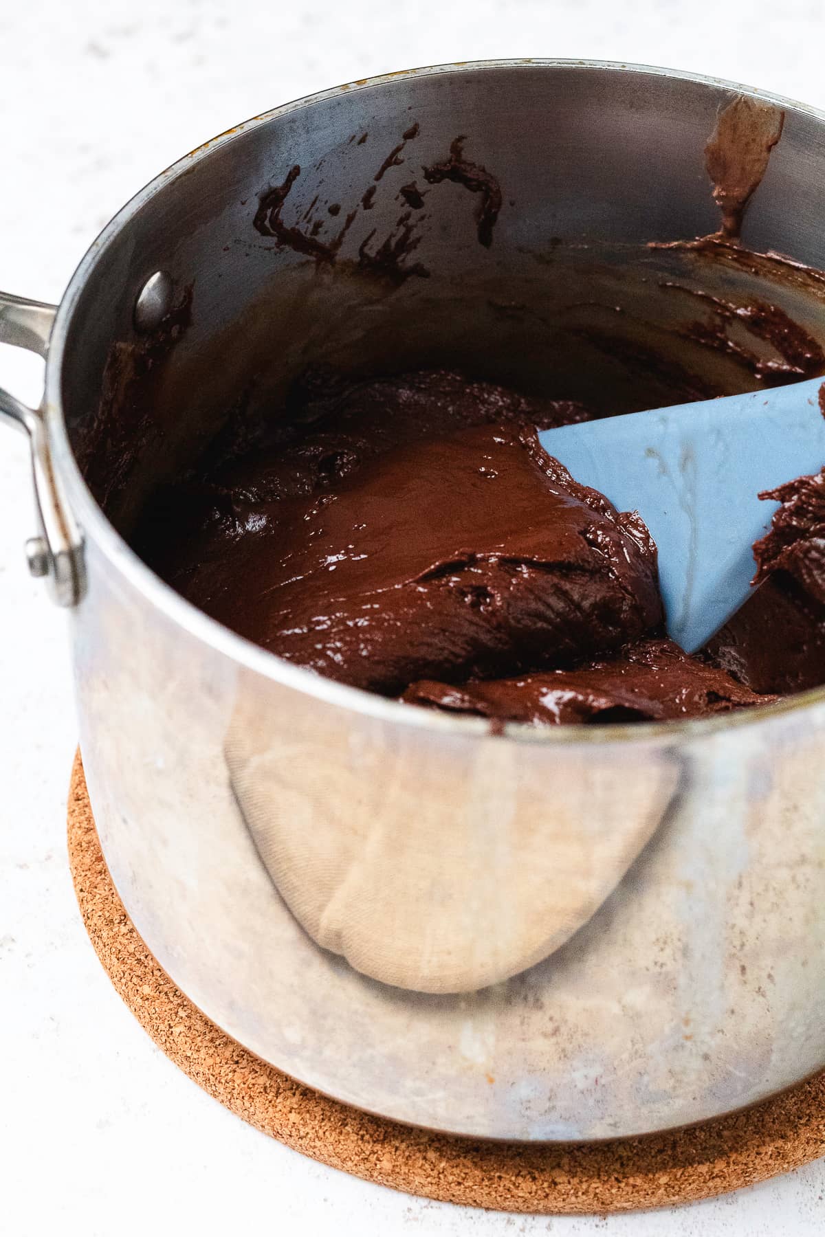Thick, melted chocolate fudge being stirred with a blue spatula in a saucepan.