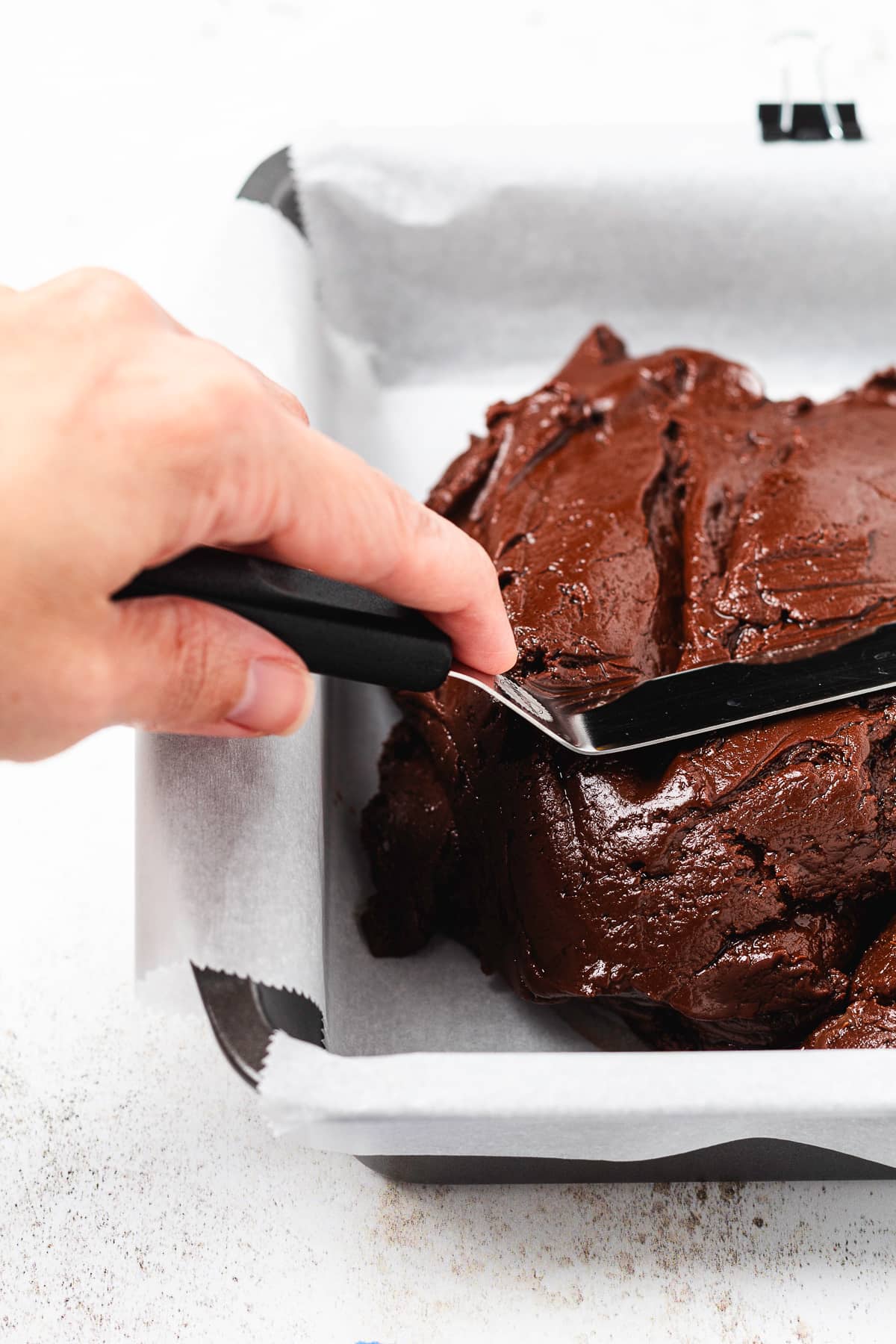 A hand spreads chocolate peppermint fudge into a parchment-lined baking pan.