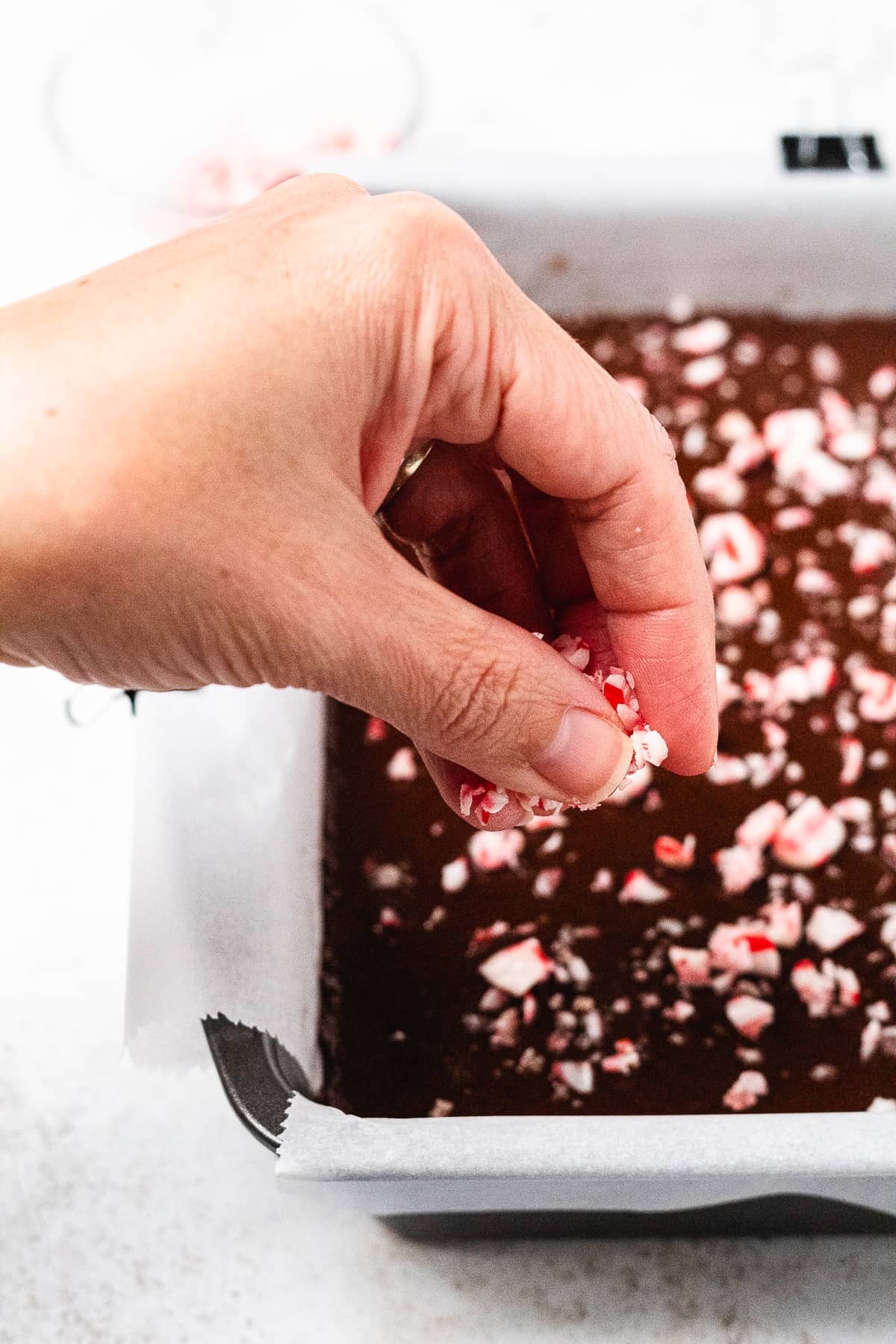 A hand sprinkles crushed candy canes over chocolate fudge in a lined baking pan.