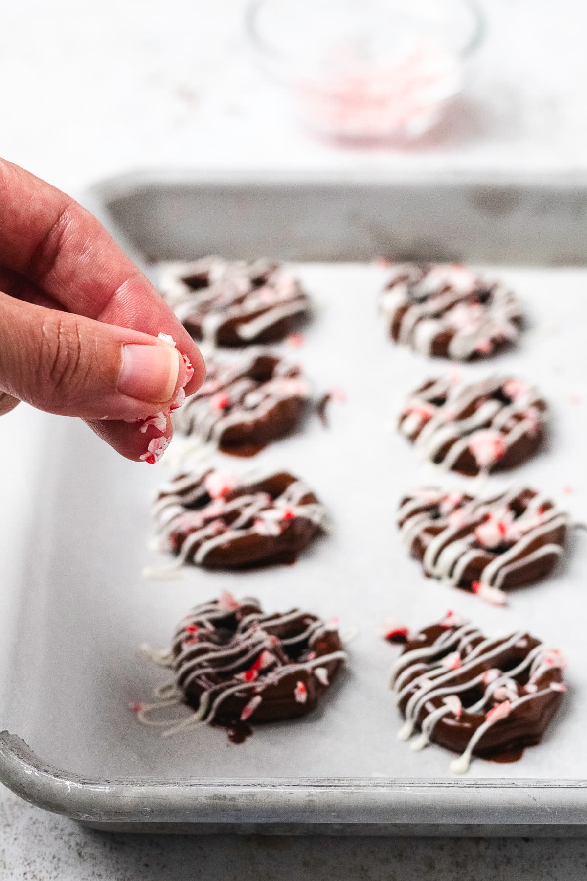 Crushed candy canes being sprinkled over chocolate-covered pretzels with white chocolate drizzle.