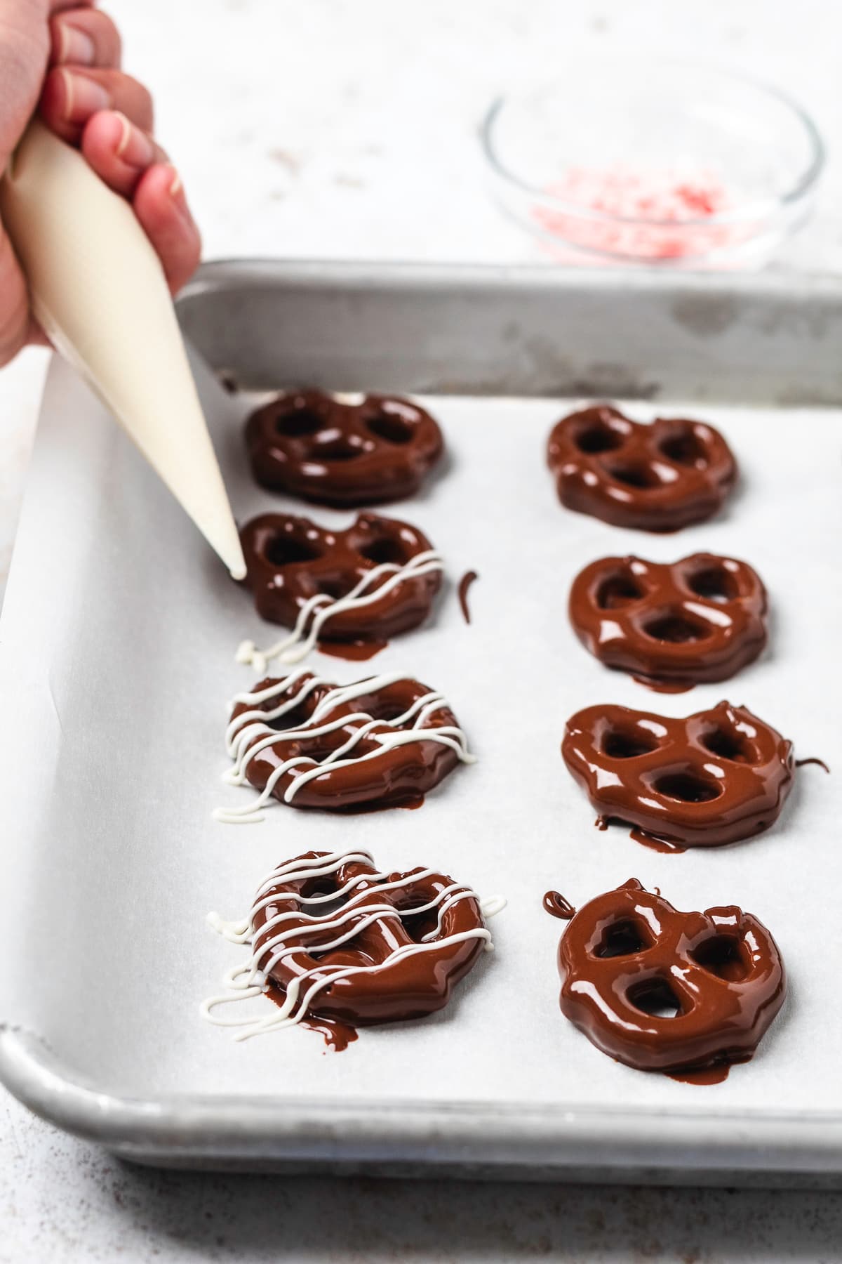Melted white chocolate being drizzled over chocolate-covered mini pretzels on a baking sheet.