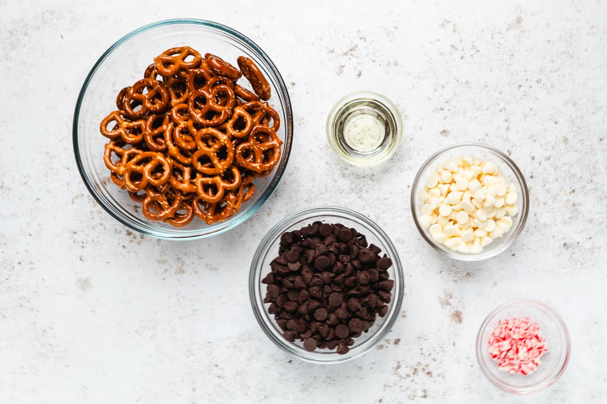 Mini pretzels, chocolate chips, white chocolate chips, oil, and crushed peppermint in glass bowls.