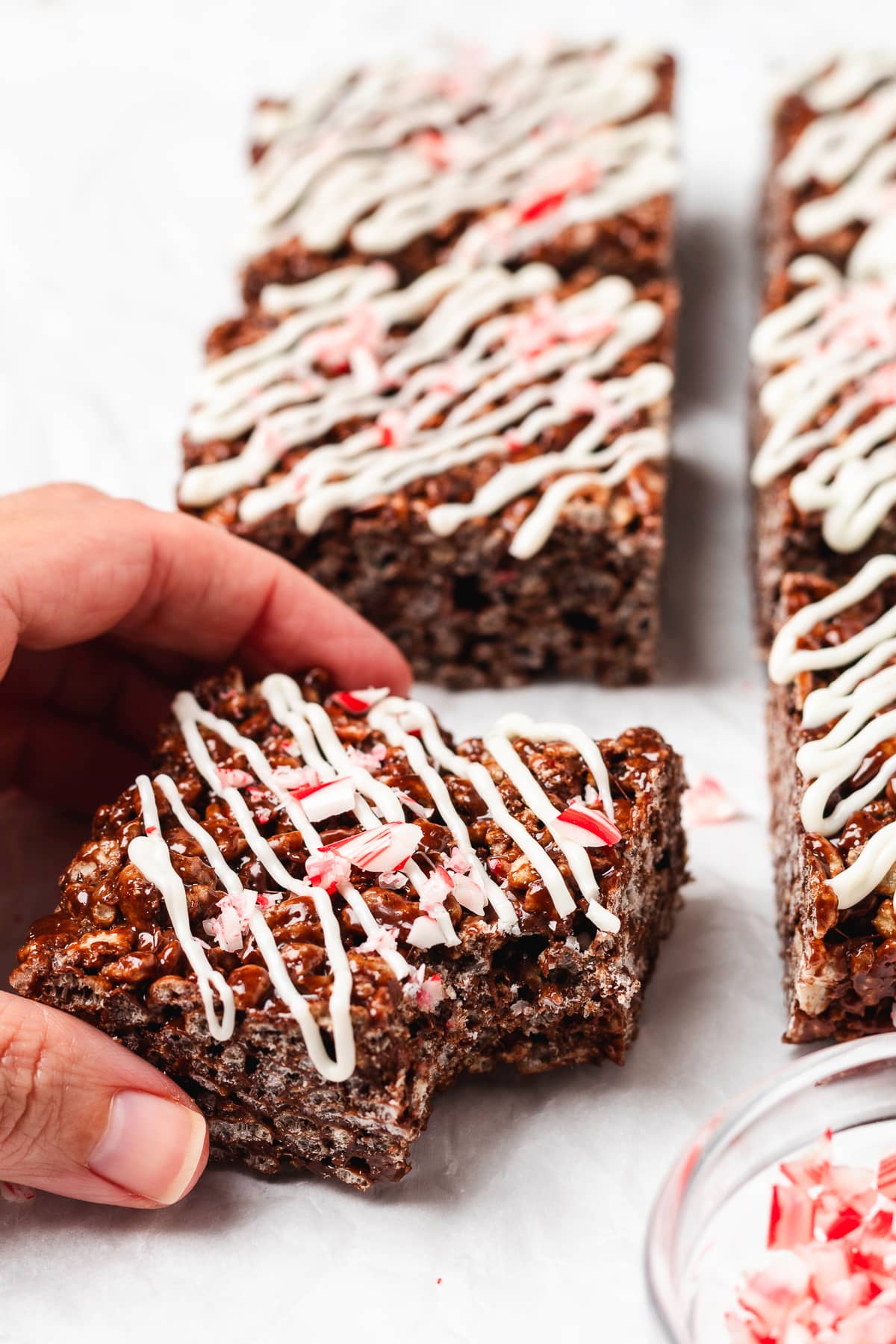 A hand picks up a chocolate peppermint Rice Krispie treat topped with white chocolate drizzle and candy canes.