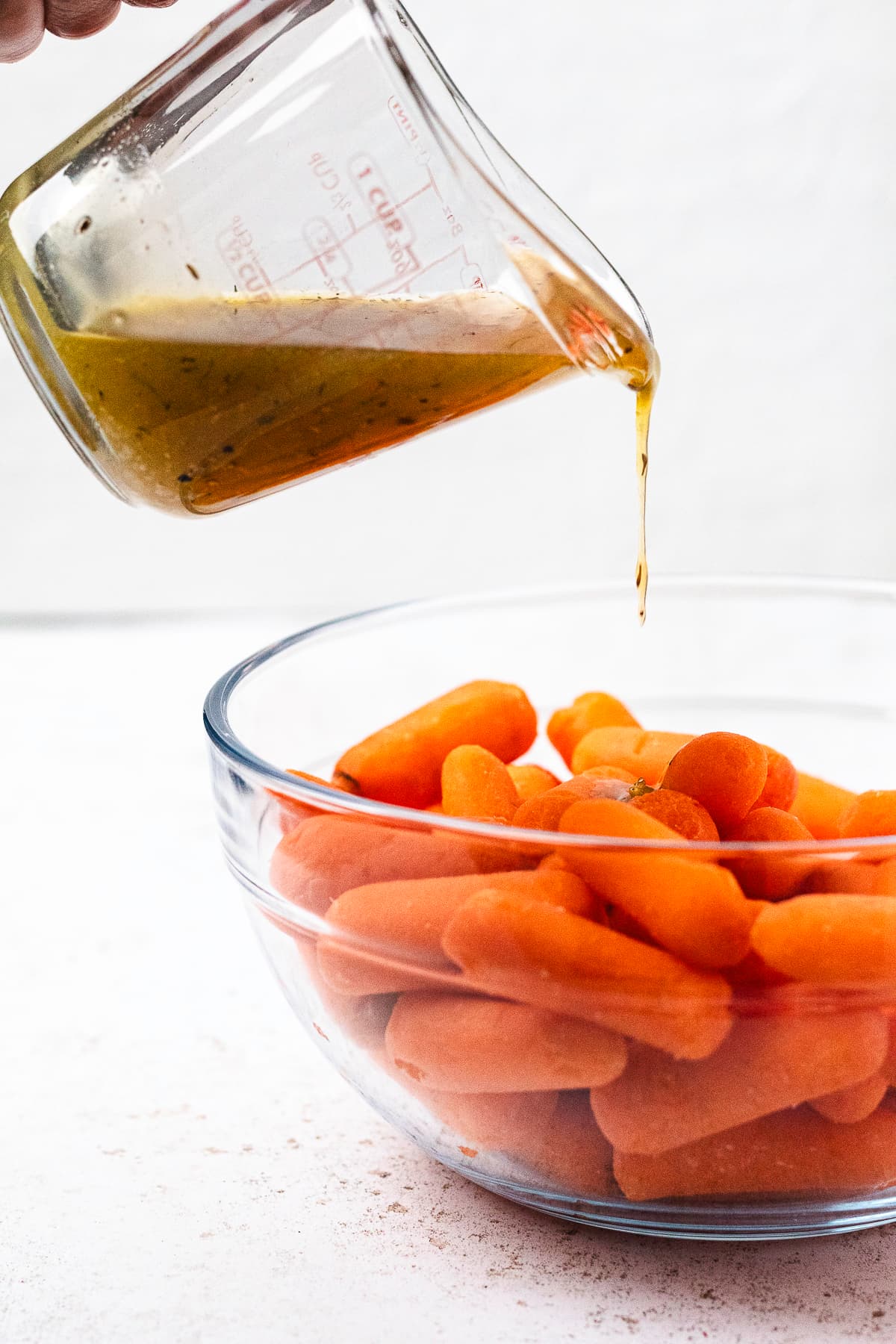 Pouring honey glaze from a measuring cup over baby carrots in a clear glass mixing bowl.