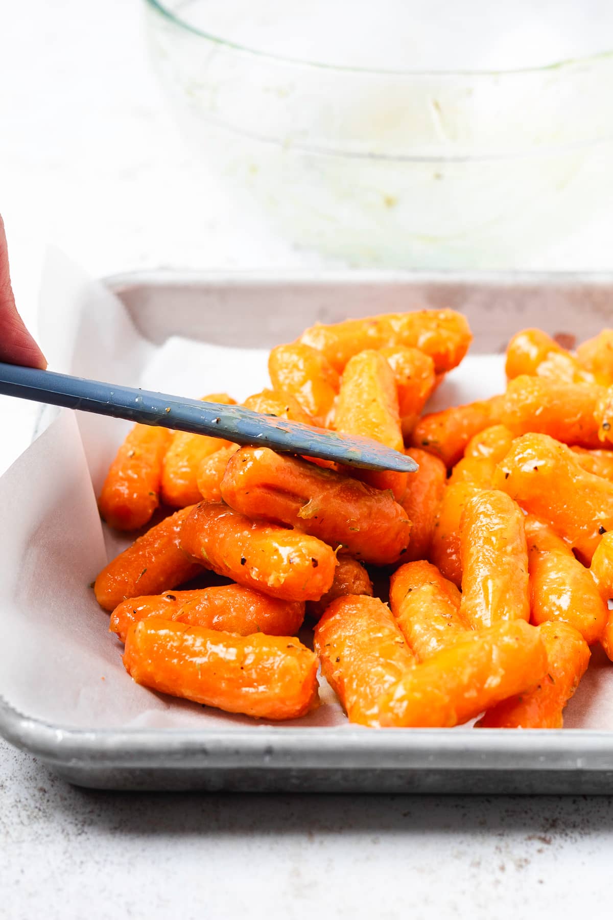 Spreading honey-glazed baby carrots on a parchment-lined baking sheet with a spatula before roasting.