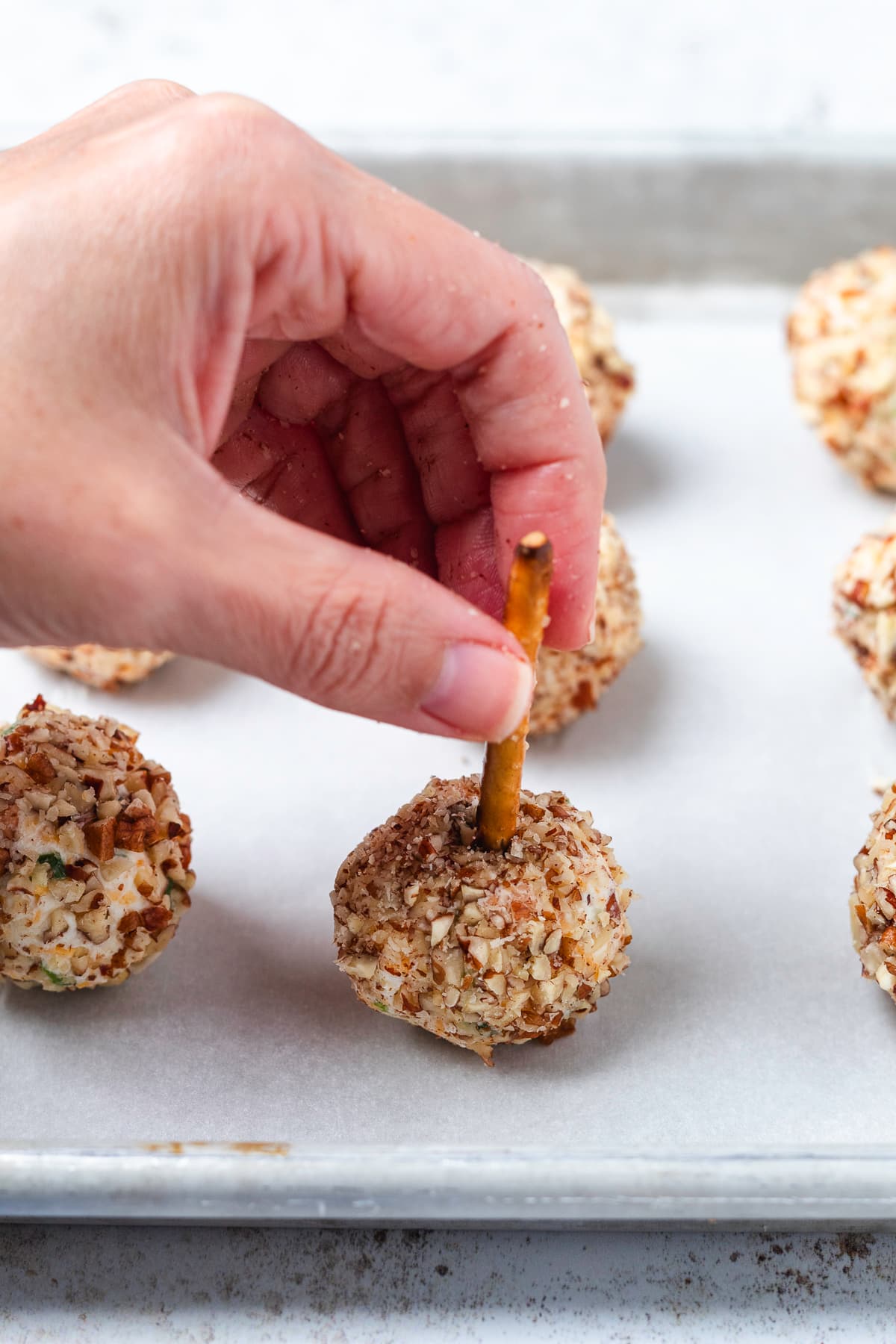 A hand inserts a pretzel stick into a pecan-coated mini cheese ball on a baking sheet.