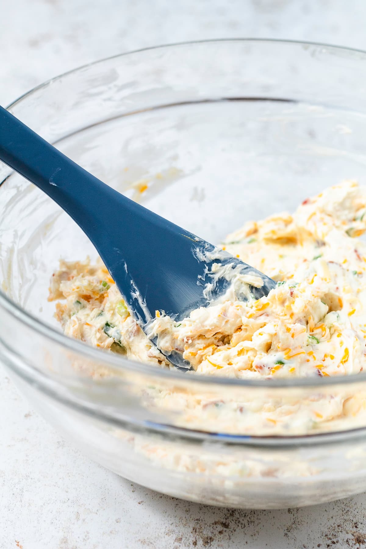 Creamy mini cheese ball mixture with shredded cheese, bacon, and green onions being mixed with a blue spatula.
