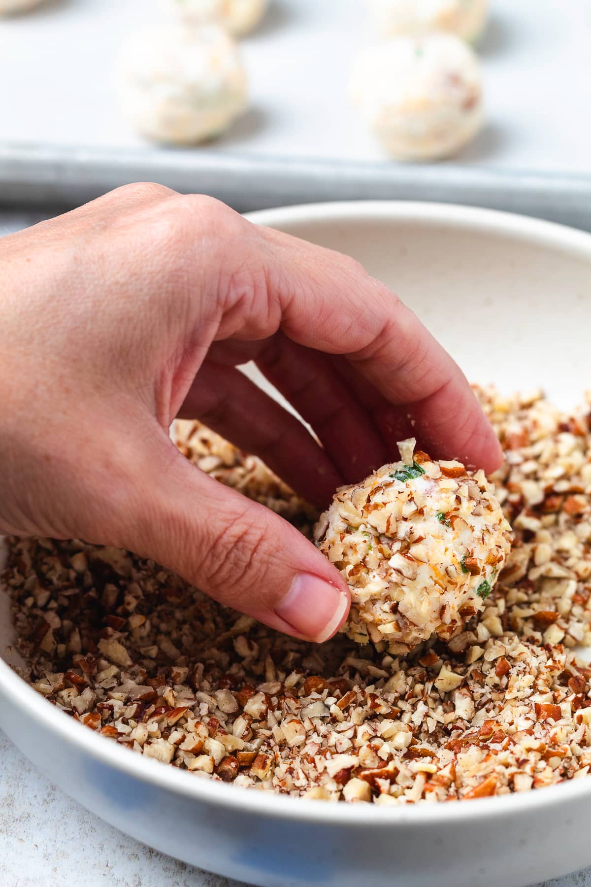 A hand rolls a mini cheese ball in finely chopped pecans to coat the outside.
