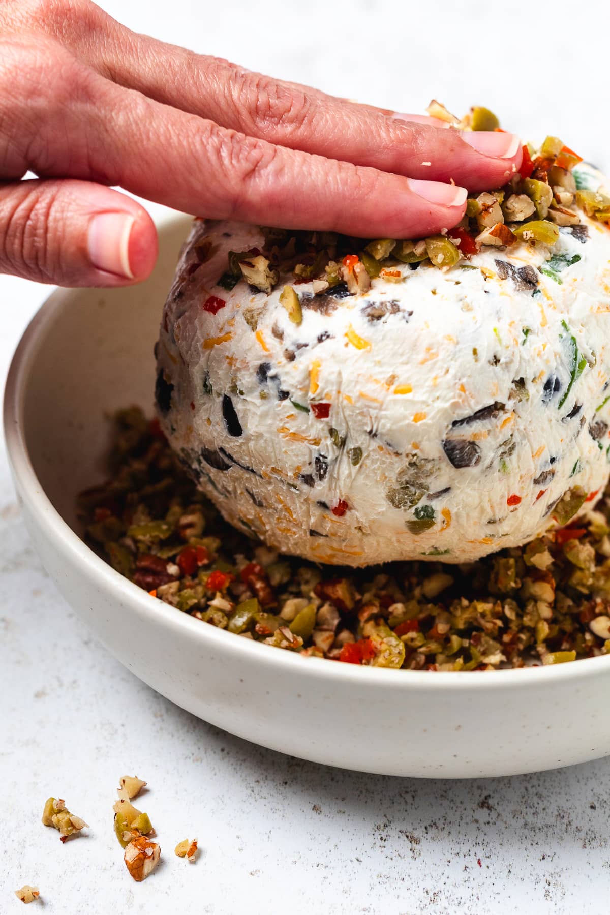 A hand presses chopped olives, pecans, and red peppers onto a cream cheese ball.