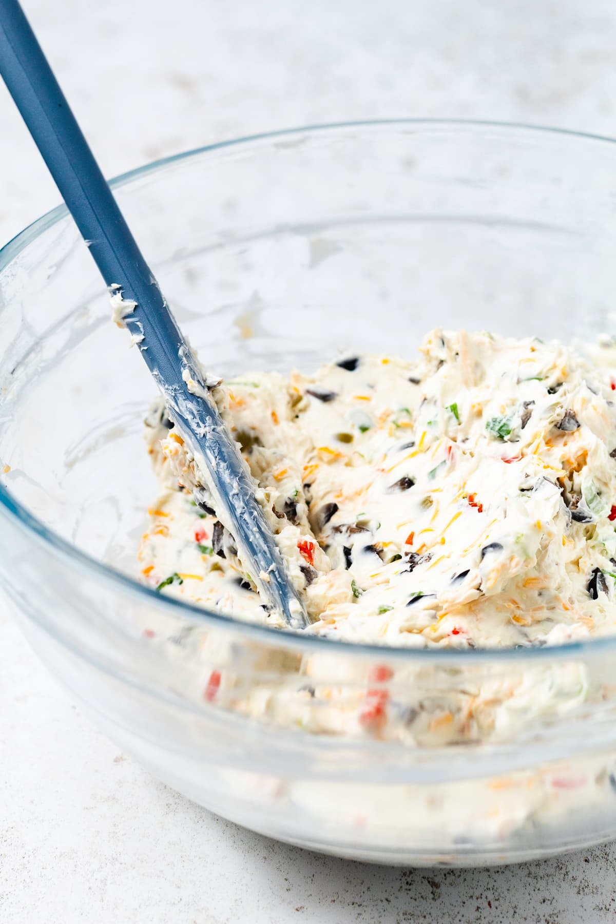 Cream cheese mixture with chopped olives, shredded cheddar, and green onions in a glass mixing bowl.