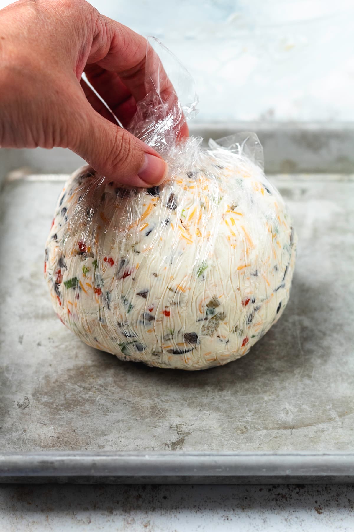 A homemade olive cheese ball wrapped tightly in plastic wrap on a baking sheet.