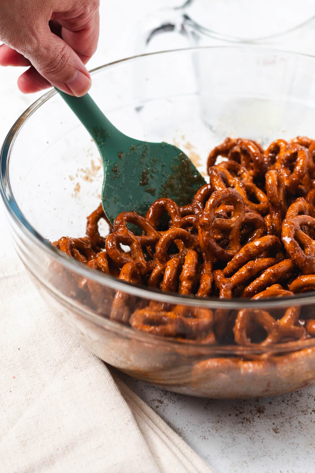 A hand uses a green spatula to mix pumpkin spice pretzels in a large glass bowl.