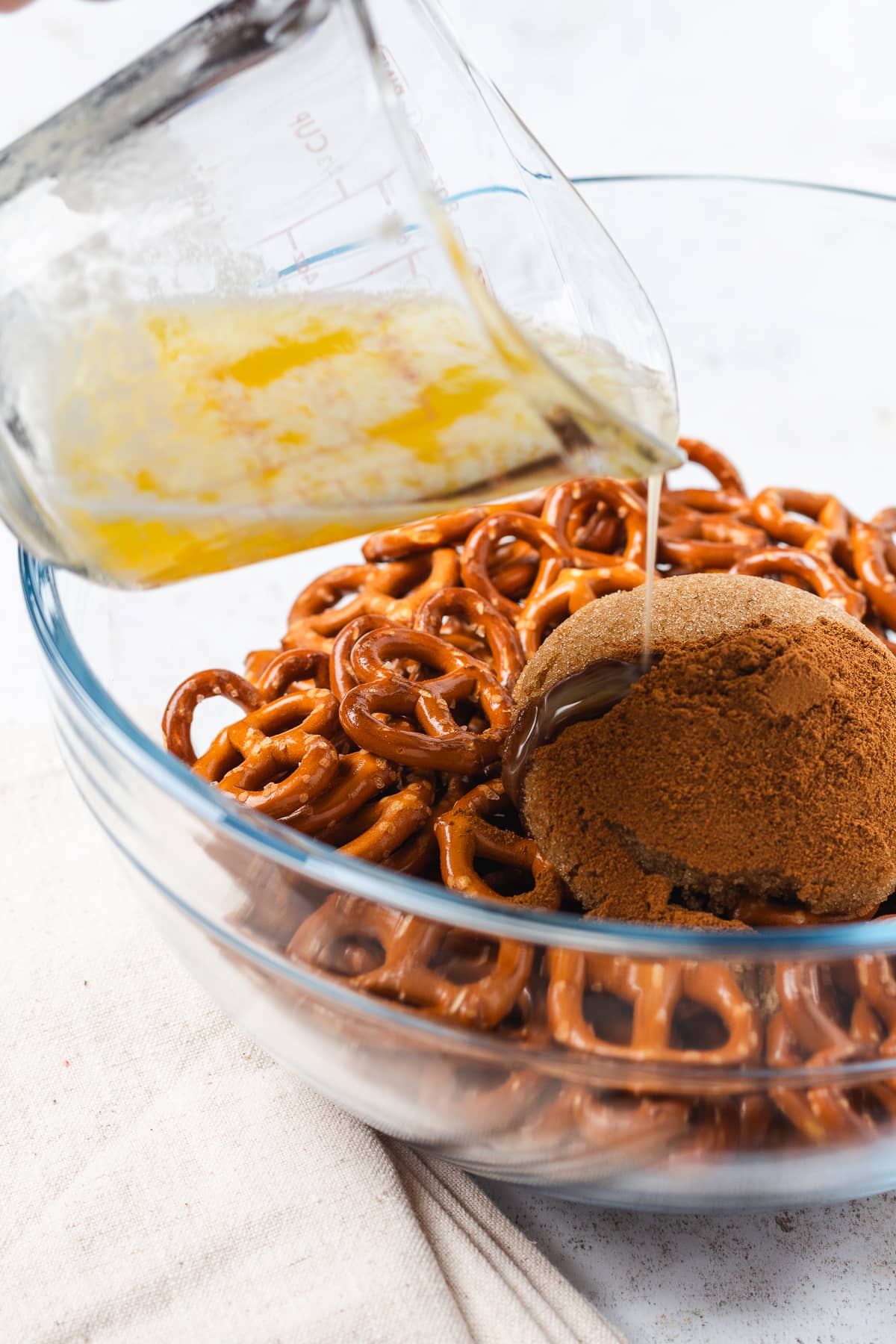 Melted butter being poured over mini pretzels, brown sugar, and pumpkin spice in a glass mixing bowl.