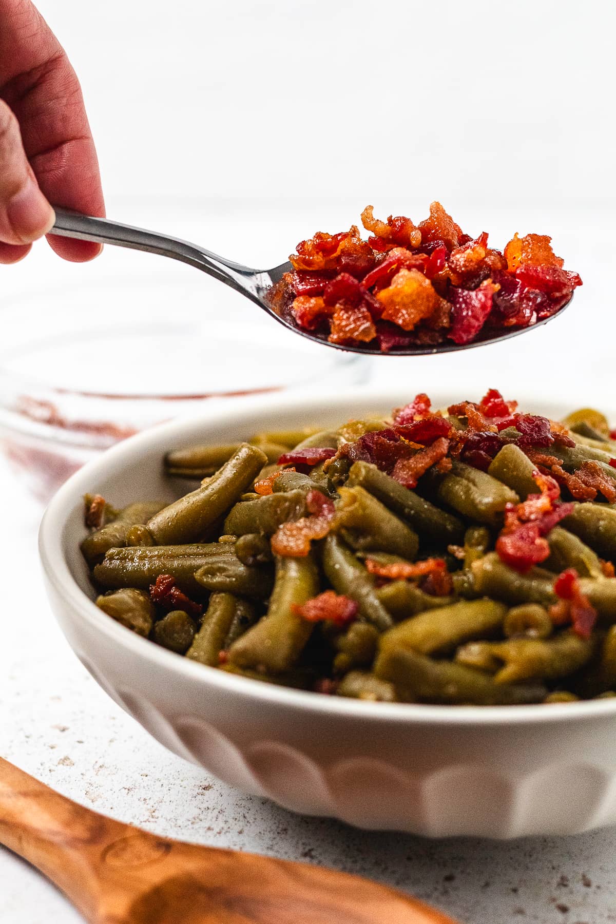 Close-up of crispy bacon on a spoon above a bowl of finished slow cooker crack green beans.